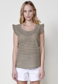 Top de ganchillo en color taupe claro con mangas cortas y volantes, que presenta un patrón de cuadrados y círculos abiertos. Combinado con pantalones cortos blancos.