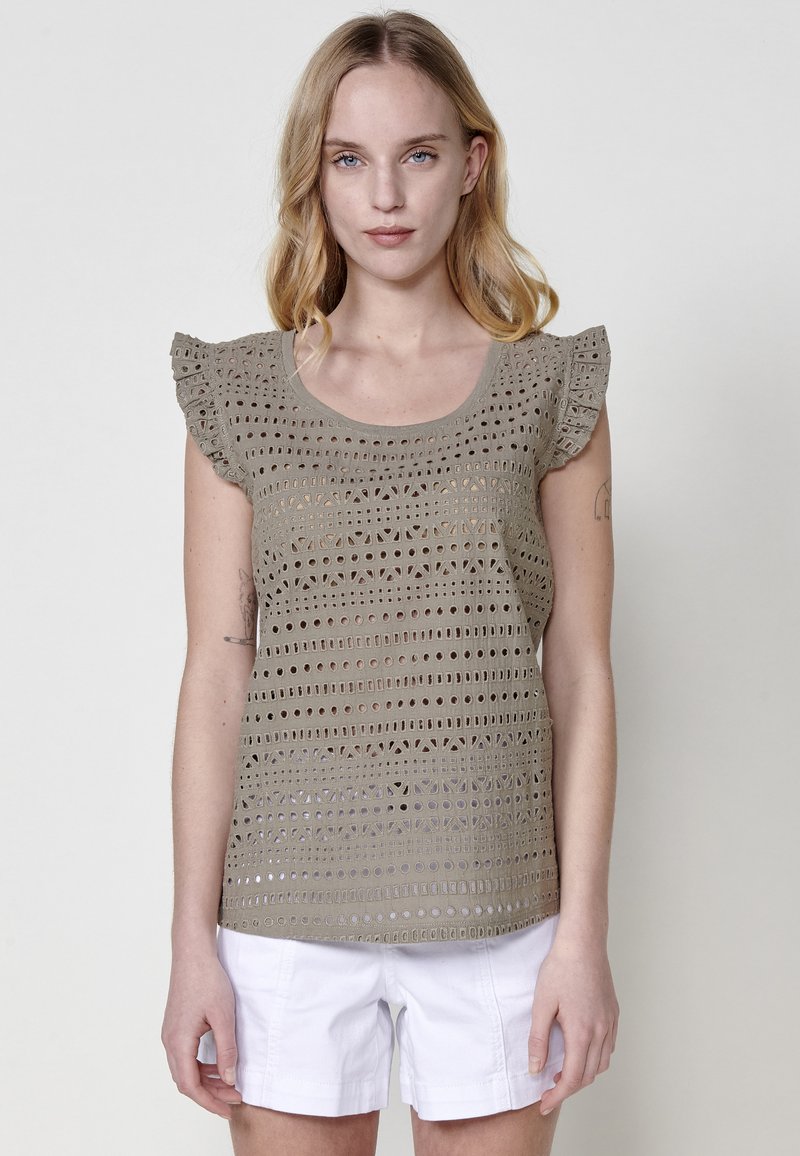 Top de ganchillo en color taupe claro con mangas cortas y volantes, que presenta un patrón de cuadrados y círculos abiertos. Combinado con pantalones cortos blancos.