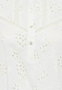 Blouse en coton blanc avec des motifs floraux brodés, des découpes, et une patte de boutonnage ornée de boutons nacrés.