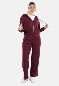 Set di felpa e pantaloni color bordeaux realizzato in tessuto morbido. La felpa presenta una chiusura con zip e cordino, maniche lunghe e una vestibilità comoda.