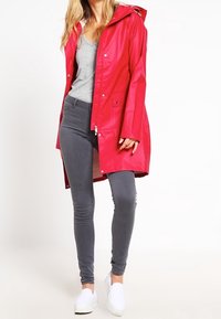 Red, waterproof raincoat with a hood, snap fasteners, and a mid-length design. Paired with gray leggings and white slip-on shoes.