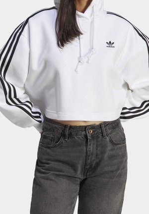 White cropped Adidas hoodie with black stripes on sleeves and logo on chest, worn with high-waisted black denim jeans.