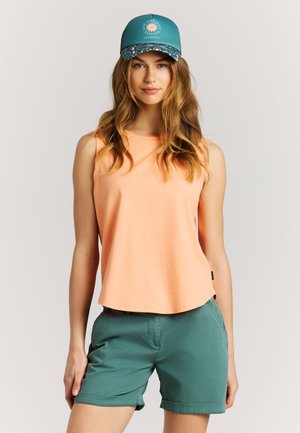 Woman wearing teal cap with sun logo, peach sleeveless top, and teal shorts, standing against a light background.