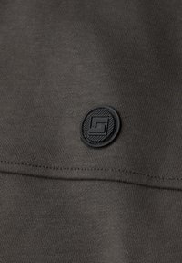 Gros plan sur un tissu gris foncé avec une couture horizontale et un petit logo circulaire noir présentant un dessin géométrique.