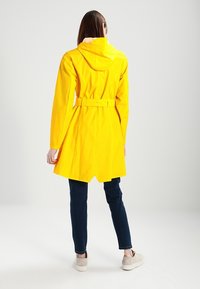 Gul vattentät trenchcoat med huva, insvept midja och en lätt utställd nederkant, kombinerad med mörka jeans och ljusa skor.