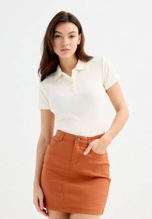 Camisa polo de canalé en color crema con mangas cortas, presenta un cuello y una botonera. Combinada con una falda de mezclilla ajustada en color óxido.
