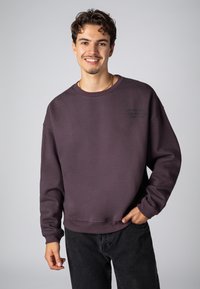 Dunkelpurple Sweatshirt mit Rundhalsausschnitt und gerippten Bündchen. Mit einer Frontaufschrift in kontrastierender Farbe. Kombiniert mit schwarzen Hosen.