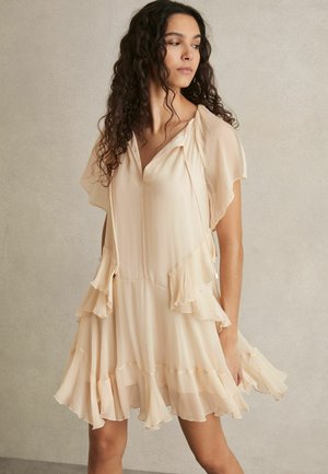 Femme aux cheveux bouclés portant une robe fluide beige clair avec des manches et un ourlet à volants, debout contre un mur uni, regardant sur le côté.