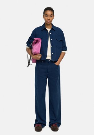Kobieta ubrana w granatową kurtkę i szerokie spodnie, trzymająca złożoną jasnoróżową torbę, stojąca z jedną ręką w kieszeni.