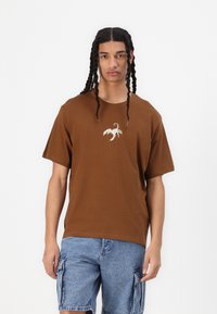 T-shirt in cotone marrone con maniche corte, caratterizzato da un design di uno scorpione bianco sul petto. Abbinato a pantaloni cargo in denim.