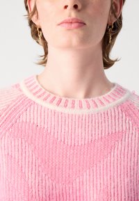 Close-up van een persoon die een roze gebreide trui met een hartpatroon en gouden hangende oorbellen draagt, op een witte achtergrond.