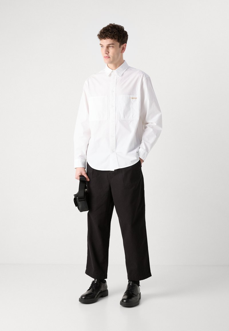 Camicia bianca a maniche lunghe con due tasche frontali, abbinata a pantaloni neri a gamba larga e scarpe nere stringate, mentre tiene in mano una borsa nera per la fotocamera.