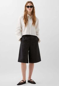 Chemise blanche à rayures, à boutons, avec épaules tombantes ; shorts noirs jusqu'aux genoux ; chaussures plates noires ; lunettes de soleil ; textures lisses.