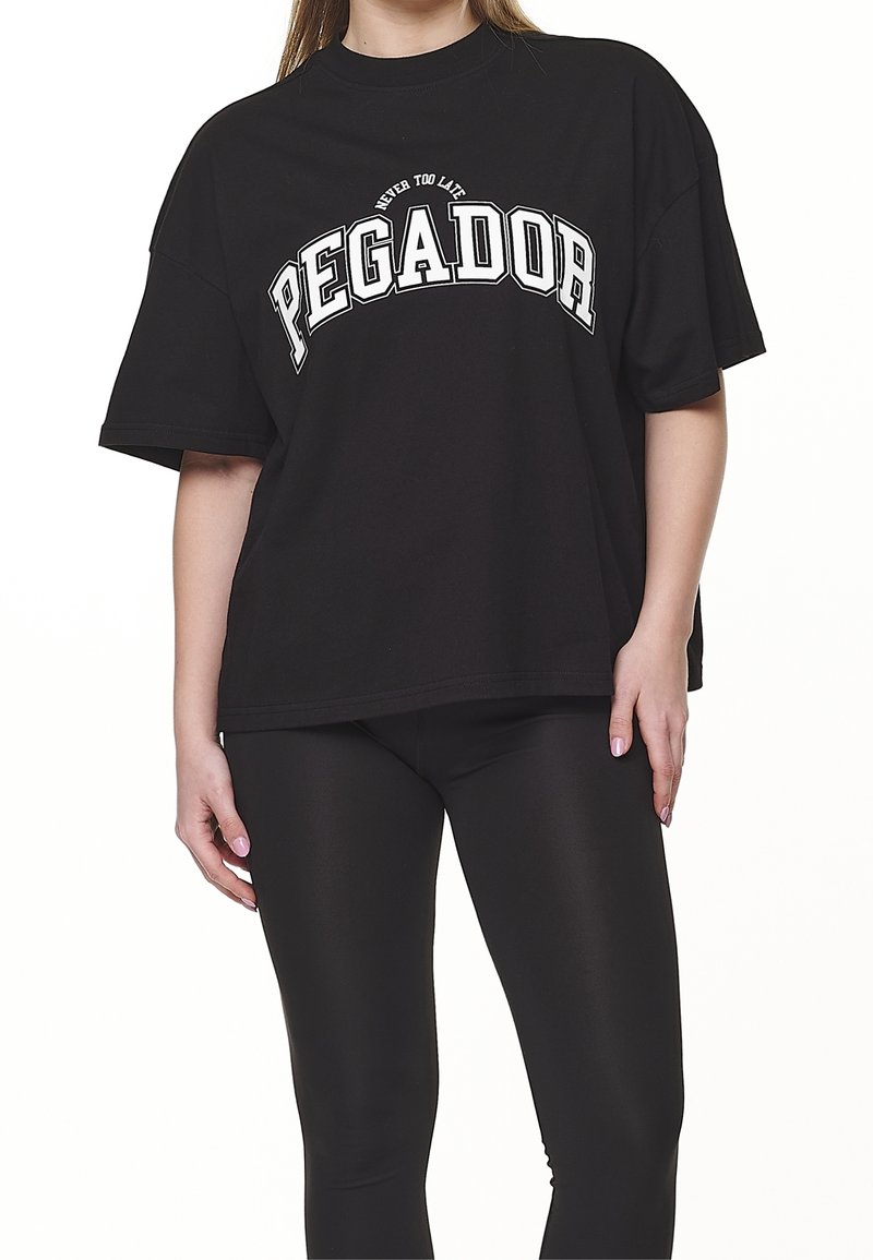 Svart oversized t-shirt i bomull, med djärv vit text som säger "PEGADOR" och frasen "ALDRIG FÖR SENT" ovanför.