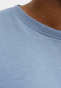 Blå bomulls-T-skjorte med rund hals, som har en jevn tekstur og et subtilt, ensartet mønster. Nærbilde av stoffet.