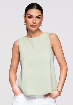 Giovane donna con capelli scuri raccolti in una coda di cavallo, che indossa orecchini a cerchio dorati, un top verde chiaro senza maniche, pantaloni bianchi e un braccialetto d’oro.