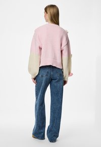Ροζ και κρεμ πλεκτή ζακέτα με μπαλονάκια μανίκια και υφασμάτινες λεπτομέρειες, συνδυασμένη με τζιν ψηλής μέσης σε μπλε χρώμα. Απλό φόντο.