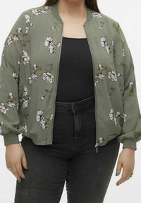 Olivengrøn bomberjakke med blomsterprint, med lynlås og ribbede manchetter. Båret over en sort top og parret med mørke jeans.