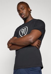 T-shirt noir à manches courtes avec un grand chiffre blanc "90" dans un design ovale sur le devant ; matière en coton doux ; coupe décontractée.