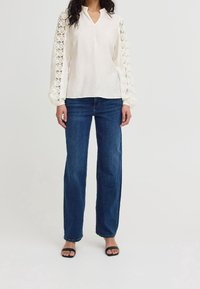 Blouse blanche à manches en dentelle, coupe décontractée et encolure en V. Associée à un jean droit bleu foncé et des sandales plates noires.