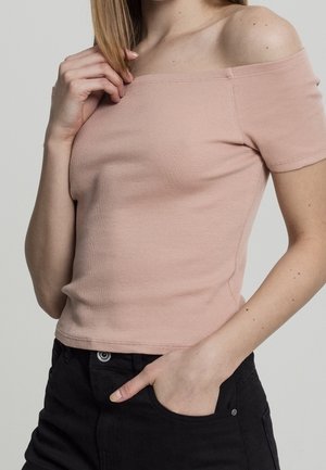 Vrouw die een lichtroze off-shoulder geribd top en een zwarte jeans draagt, met de ene hand haar sleutelbeen aanrakend en de andere in haar zak.