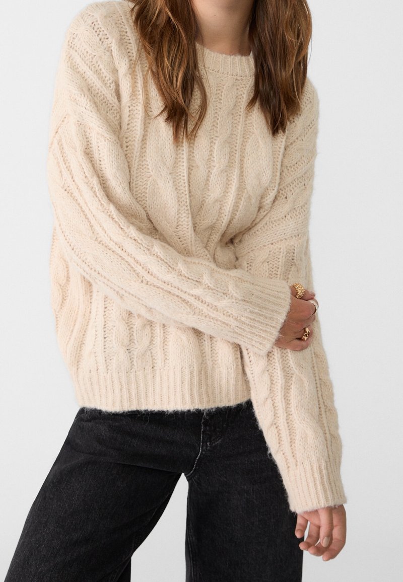 Femme portant un pull torsadé crème à manches longues et un jean noir, tenant une manche avec sa main ornée de bagues en or.