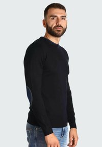 Pull en tricot noir avec un col rond, présentant un accent bleu foncé au niveau des coudes. Le modèle le porte avec un jean en denim bleu clair.