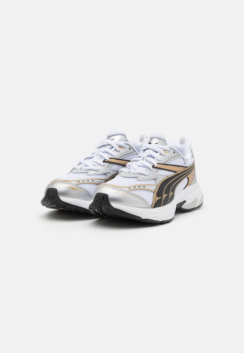 Zapatillas deportivas blancas con detalles en plata y negro, parte superior de malla y suela de goma texturizada. Presenta cordones y elementos de diseño laterales distintivos.