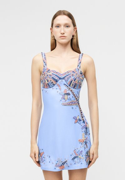 Jeune femme portant une mini robe bleu clair ornée de motifs d'étoiles de mer et de coquillages en perles, debout devant un fond blanc uni.