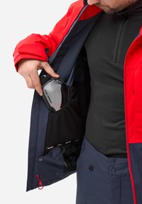Rote und marineblaue Jacke mit einer Mesh-Tasche, in der Skibrillen aufbewahrt werden. Texturiertes schwarzes Basislager sichtbar unter der Jacke.
