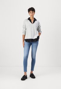 Maglione grigio chiaro a scollo a V indossato sopra una camicia nera con colletto e pois bianchi, abbinato a jeans aderenti blu e mocassini neri.