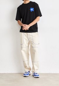 Camiseta de algodón negra con gráfico de flor azul, pantalones color crema con costuras azules, zapatillas azules y blancas, de pie sobre un suelo de concreto.