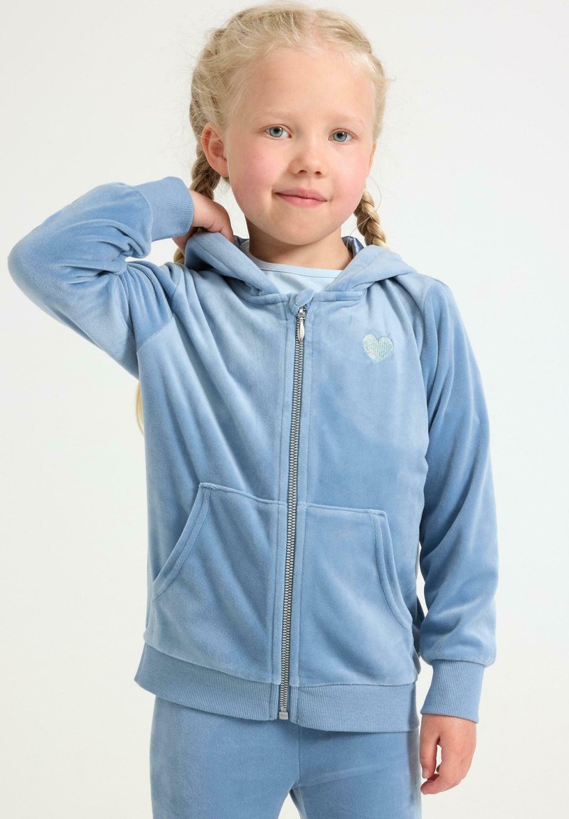 Lyseblå hætte-sweatshirt med lynlås i blødt materiale, med forlommer og et lille hjerteemblem på brystet. Glat tekstur.