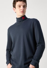 Maglione blu navy a collo alto con maniche lunghe, realizzato in un tessuto liscio, con un piccolo etichetta rossa sul colletto.