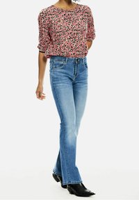 Kobieta w jasnoniebieskich dzwonowych dżinsach, czarnych butach na obcasie z ostrym noskiem oraz różowej bluzce w kwiaty z rękawami do łokcia.