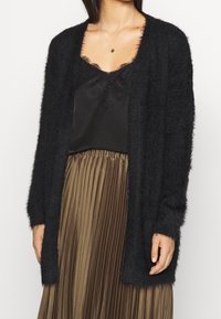 Schwarzer, flauschiger Cardigan mit offenem Frontdesign, langen Ärmeln und weicher Textur, getragen über einem schwarzen, mit Spitze besetzten Oberteil und einem plissierten braunen Rock.