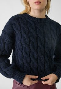Maglione lavorato a maglia di colore blu navy con motivo a trecce, caratterizzato da polsini e orlo a coste. Il tessuto texturizzato e morbido aggiunge profondità al design.