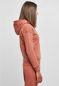 Rosa hettegenser med løs passform, som har hette og ribbestrikkede mansjetter. Laget av mykt stoff, med en glatt tekstur og minimalistisk design.