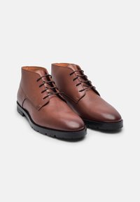 Bottines en cuir marron avec une texture lisse, bout rond et lacets marron. Semelle en caoutchouc noir avec des détails de grip et un design minimaliste.