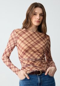 Langsleeve, pasvormtop van doorschijnende stof met een tan en bordeaux ruitpatroon. Beschikt over een ronde halslijn en geplooide details aan de zijkanten.