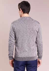 Man som står med ryggen mot kameran, iklädd en grå stickad tröja med långa ärmar och mörkblå byxor, mot en neutral bakgrund.