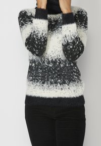 Person in einem flauschigen schwarz-weißen gemusterten Pullover und schwarzen Hosen, die teilweise ihr Gesicht mit den Händen bedecken.