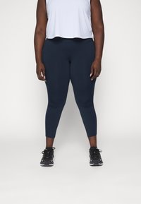 Leggings azul marino hechos de material elástico, con una cintura de media altura y longitud hasta el tobillo. Combinados con una camiseta corta gris clara y zapatillas negras.