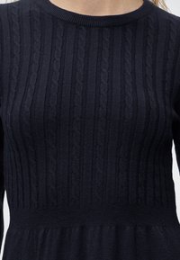 Gros plan d'un pull en maille torsadée bleu marine à col rond porté par une personne, montrant la texture et les détails du motif tricoté.