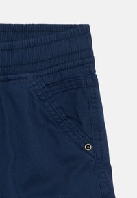 Pantalon bleu marine avec une taille élastique, poche latérale avec un bouton-pression argenté et texture en fines côtes au niveau de la taille.