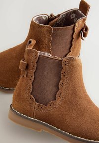 Botas de tobillo de ante marrón con paneles laterales elásticos, bordes ondulados y un interior texturizado. Presenta una suela de goma marrón y costuras sutiles.
