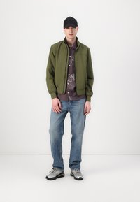 Zelena bomber jakna, siva košulja s uzorkom, plave traperice i sive tenisice. Model nosi crnu kapu, prikazujući ležeran stil.