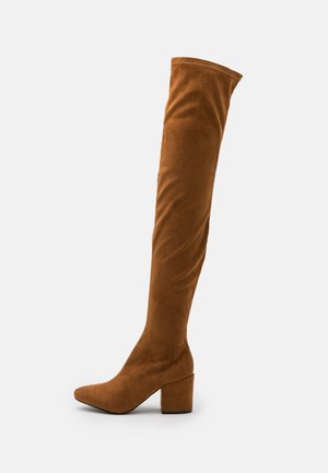 Botte haute en daim marron avec talon bloc et bout pointu, s'étendant au-dessus du genou sur un fond clair uni.