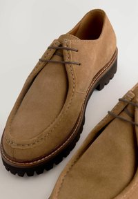 Chaussures en daim marron avec un bout rond, des détails cousus et des lacets gris. Semelle extérieure noire épaisse avec des motifs de traction pour l'accroche.