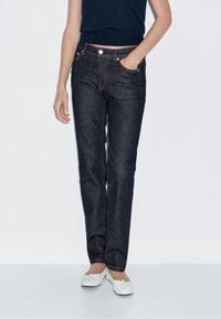 Ciemnoniebieskie dżinsy o prostym kroju, z pięcioma kieszeniami i metalowym guzikiem, zestawione z dopasowaną czarną bluzką i białymi płaskimi butami.